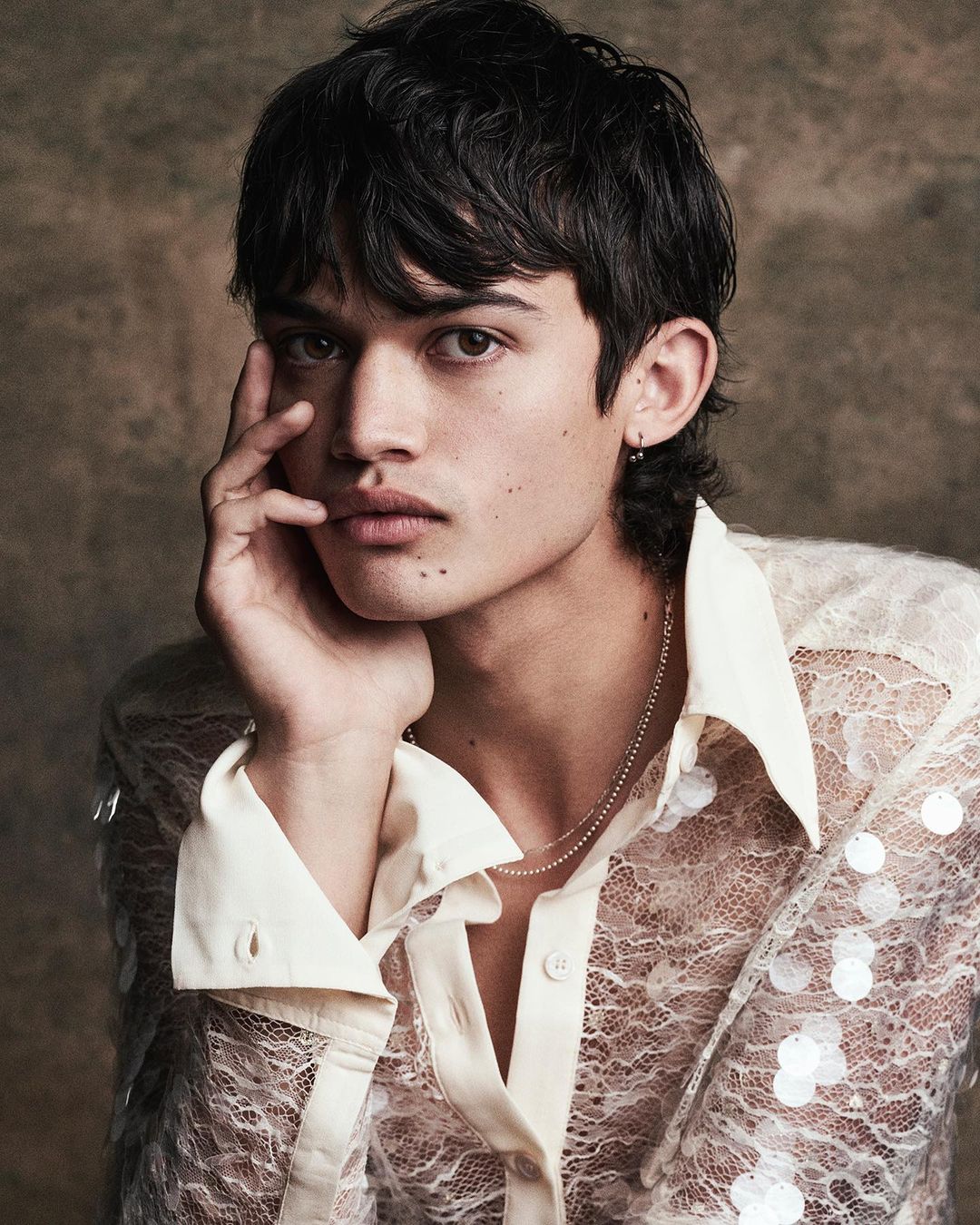 Portrait of a young adult male with dark hair and light skin, wearing a translucent, collared lace shirt in a studio setting.