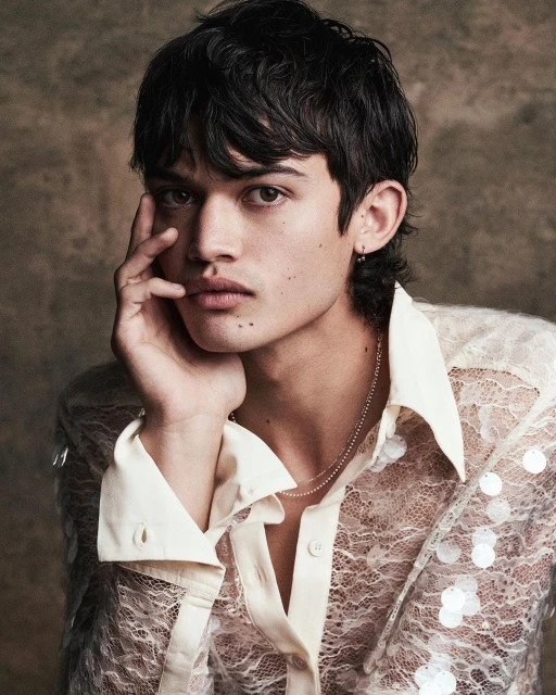 Portrait of a young adult male with dark hair and light skin, wearing a translucent, collared lace shirt in a studio setting.