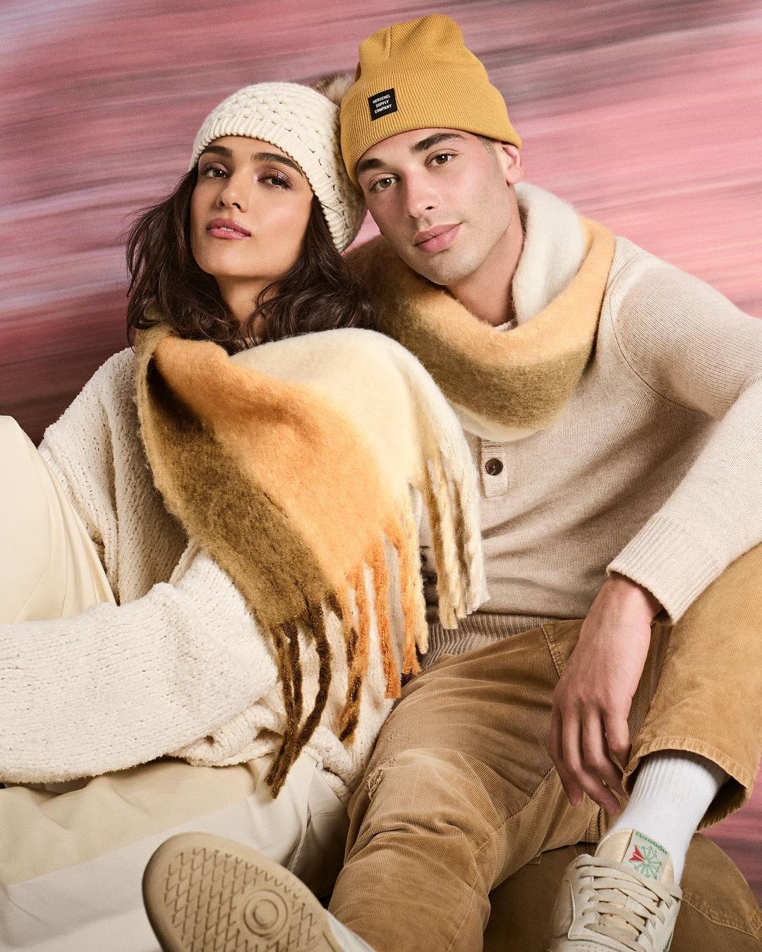 Two young adults, a male and female, wearing winter hats and scarves in a studio setting, showcasing seasonal fashion apparel.
