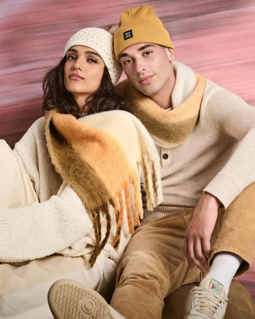 Two young adults, a male and female, wearing winter hats and scarves in a studio setting, showcasing seasonal fashion apparel.