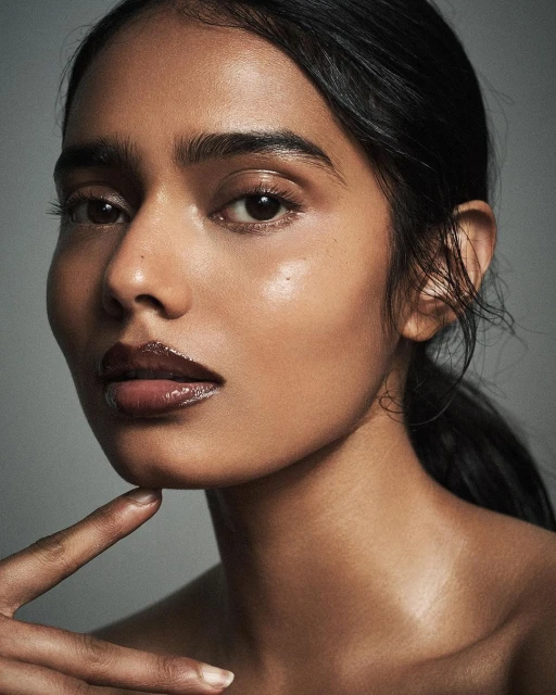 Close-up beauty shot of a woman with brown skin, dark hair pulled back, and glossy lips posing indoors against a grey background.
