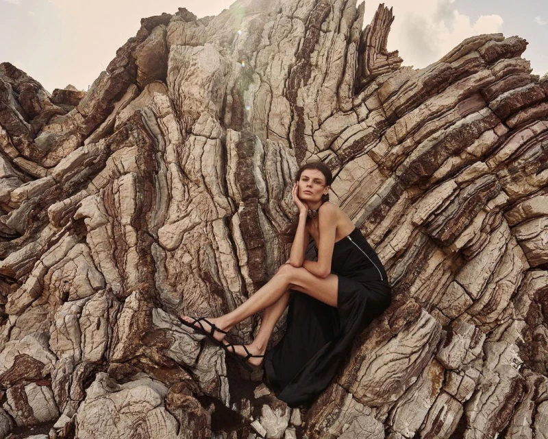 Elegant adult woman in black dress poses seated on striking layered rock formation, creating a visually compelling fashion image.