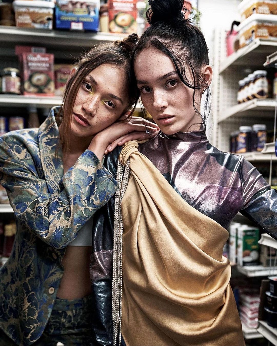 Two adult women in a store aisle pose in fashionable metallic clothing with patterned blazers and draped fabric, captured in medium shot.