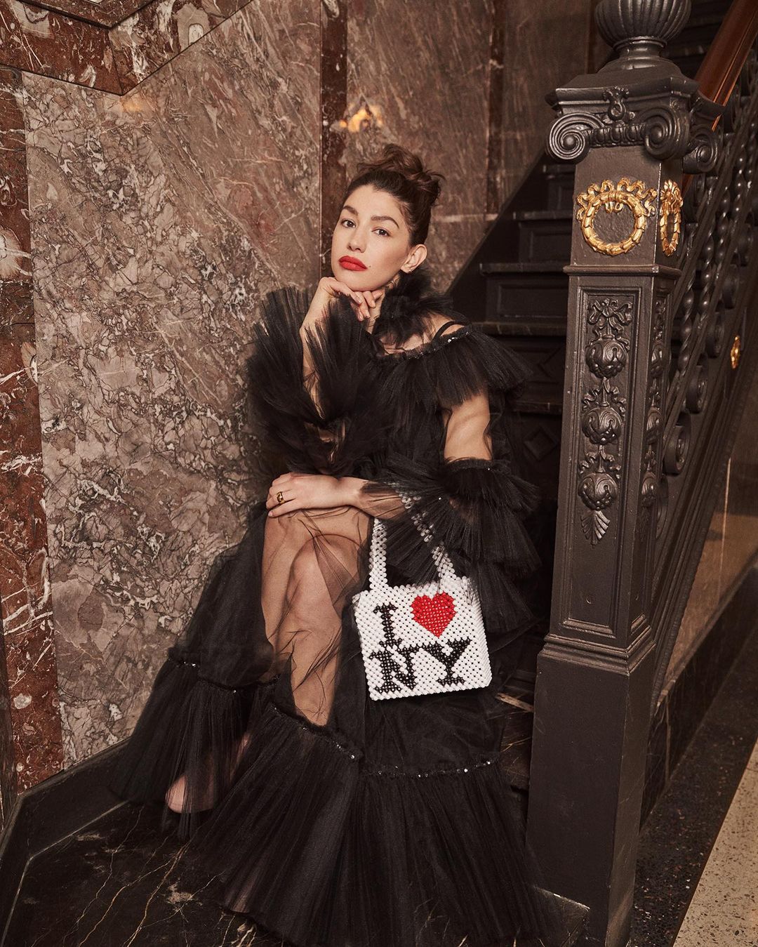 Elegant woman in a formal black dress poses on stairs with an I ❤️ NY handbag, highlighted by marble details and soft lighting.