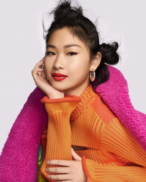 Portrait of an Asian female with a stylish hairstyle and wearing red lipstick, a ribbed top, and a pink faux fur jacket, indoors.