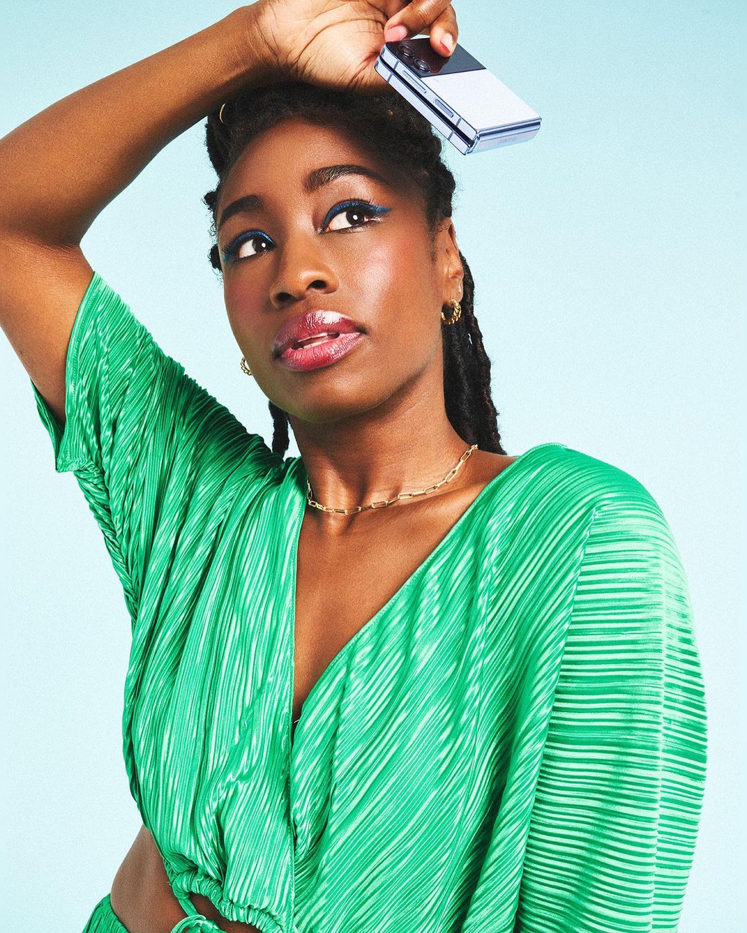 A young Black woman poses indoors with a folding smartphone above her head, wearing a green top against a light blue background.