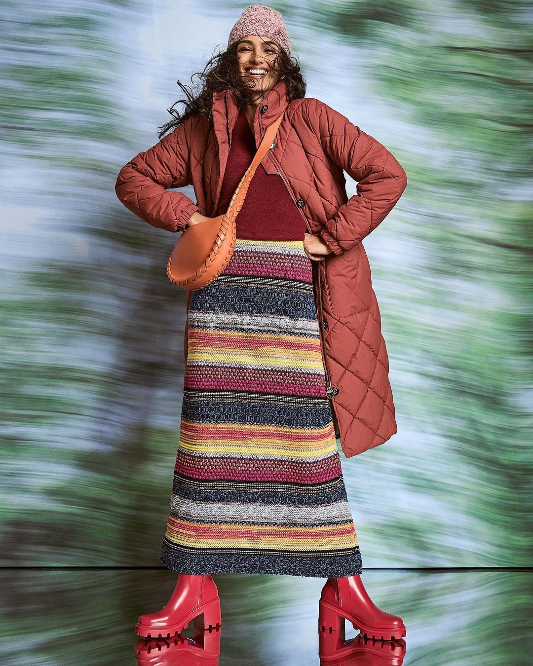 Full-length studio shot of a smiling woman wearing a long quilted jacket, knit hat, colorful striped skirt, and high-heeled boots.
