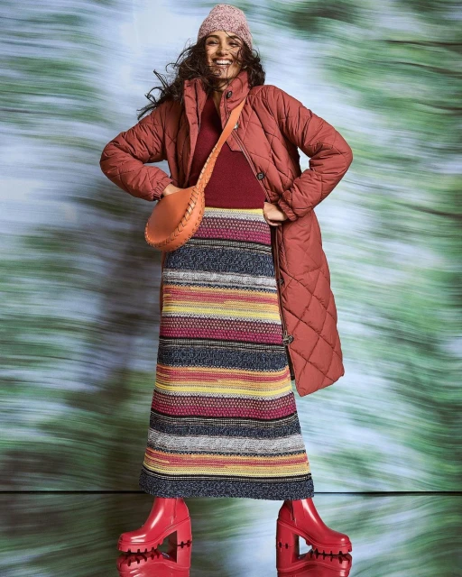 Full-length studio shot of a smiling woman wearing a long quilted jacket, knit hat, colorful striped skirt, and high-heeled boots.