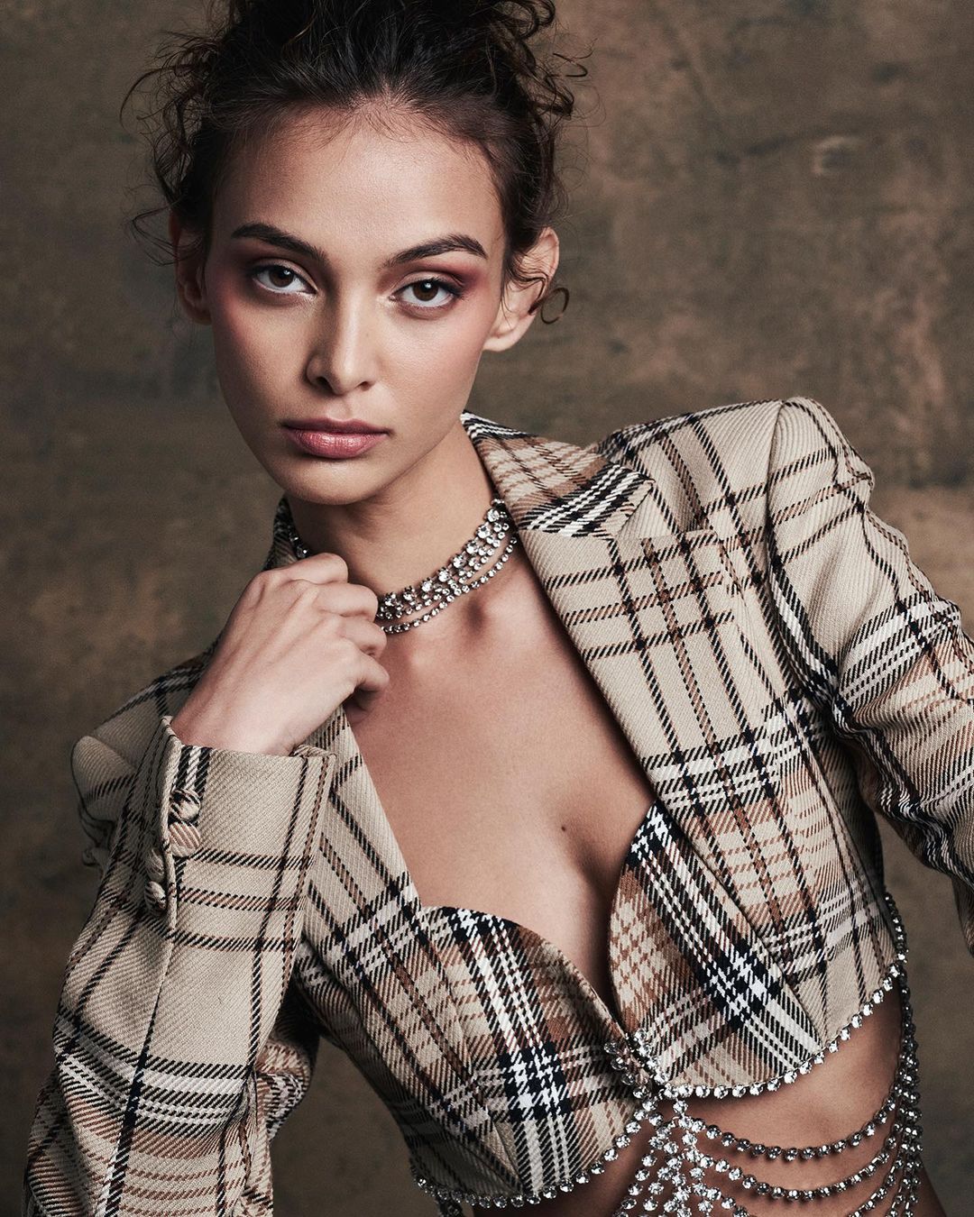Portrait of a young adult female model in a tan plaid blazer and jewel-encrusted necklace in a studio setting with soft lighting.