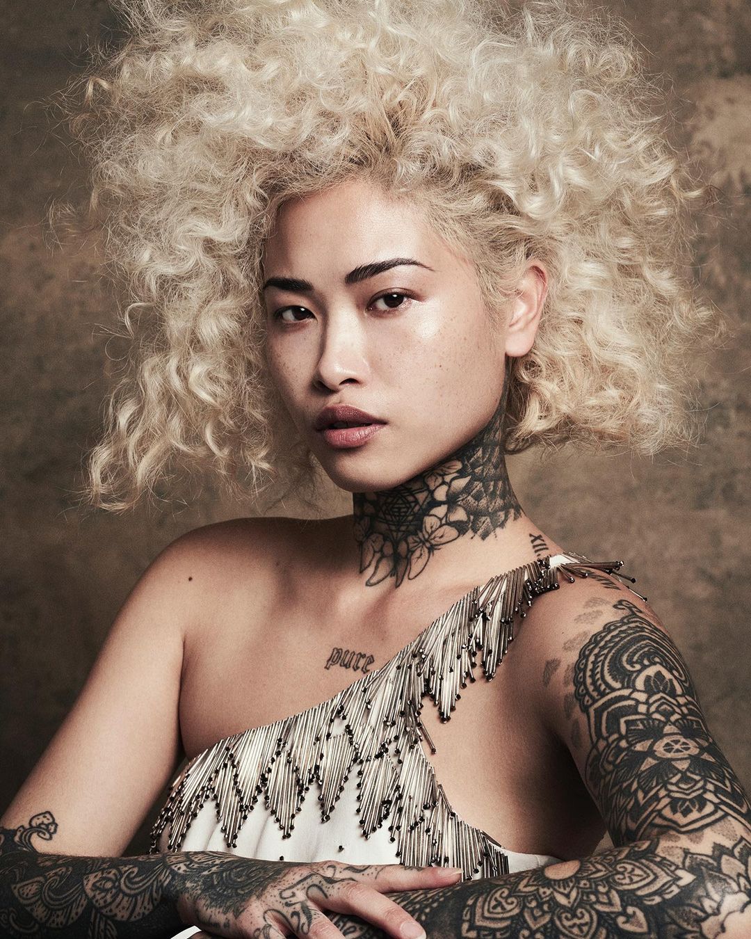 Close-up studio portrait of a young adult Asian woman with fair skin, curly blonde hair, and dark tattoos, wearing a one-shoulder dress.