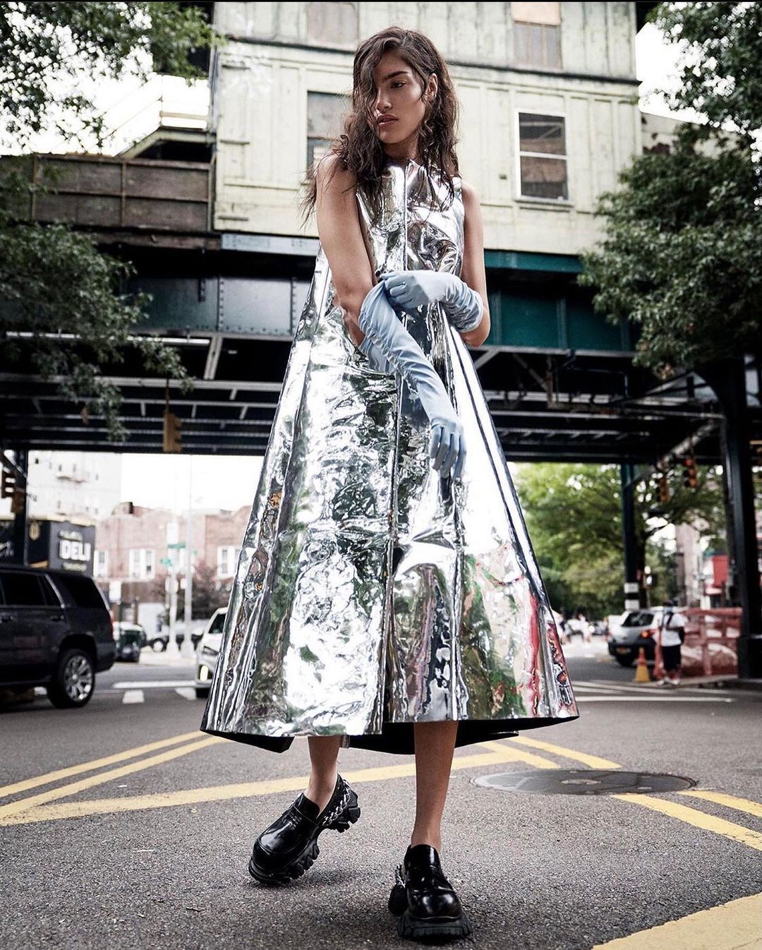 Fashion model poses in a metallic dress and long blue gloves on a city street with buildings and an elevated train overpass.