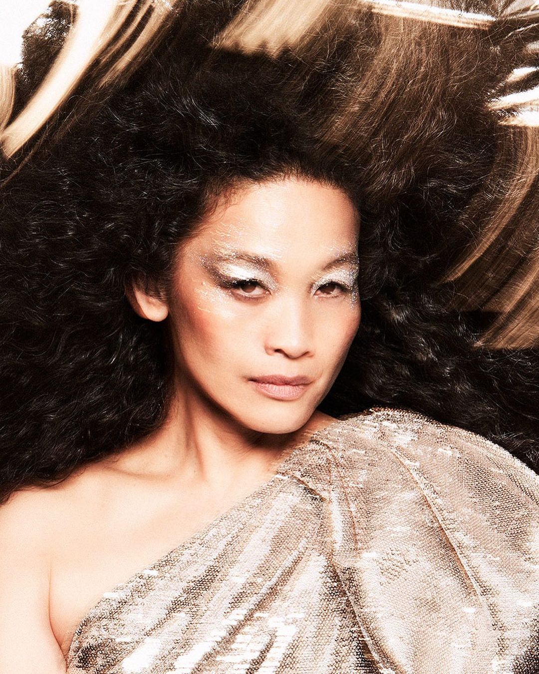 Portrait of an adult woman with dark curly hair and glittery makeup, wearing a silver sequin one-shoulder top, posing in a studio.