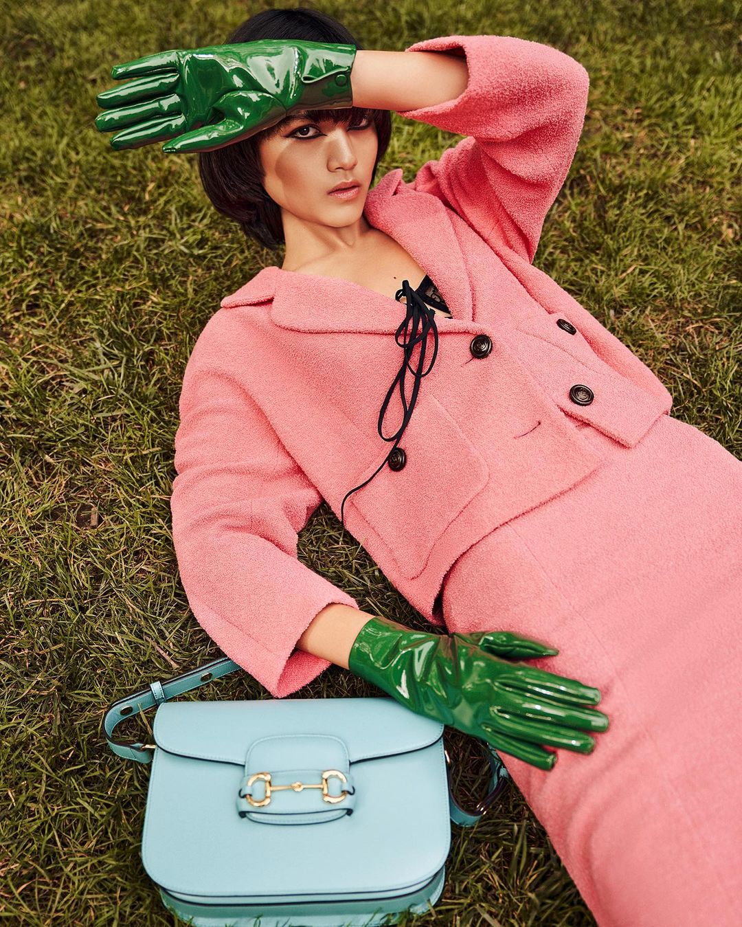 Woman lying on grass in a pink tweed suit, accented with green gloves and a blue purse, outdoor fashion shot, portrait orientation.