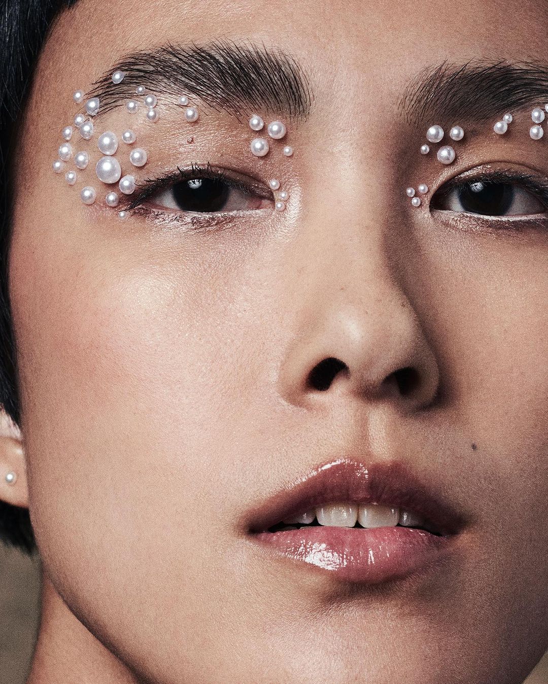 Close-up beauty shot of a woman with pearls applied as decorative makeup around her eyes and glossy lips; neutral skin tone and background.