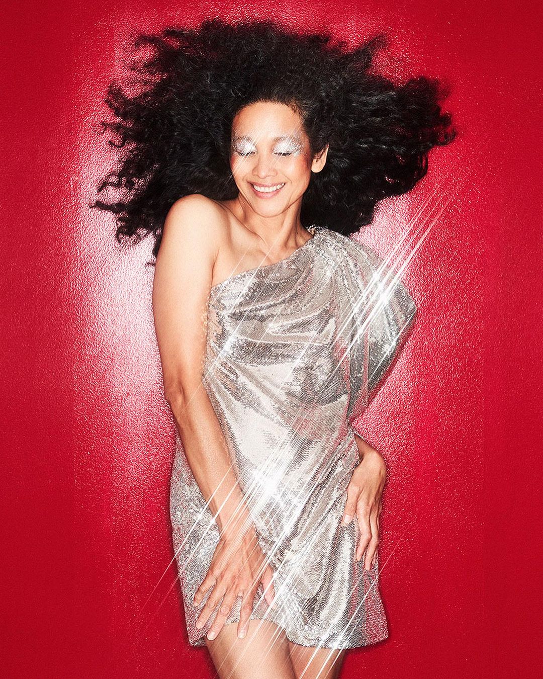 Smiling adult female with curly dark hair and shimmering silver eye makeup, wearing a shiny silver dress against a textured red background.