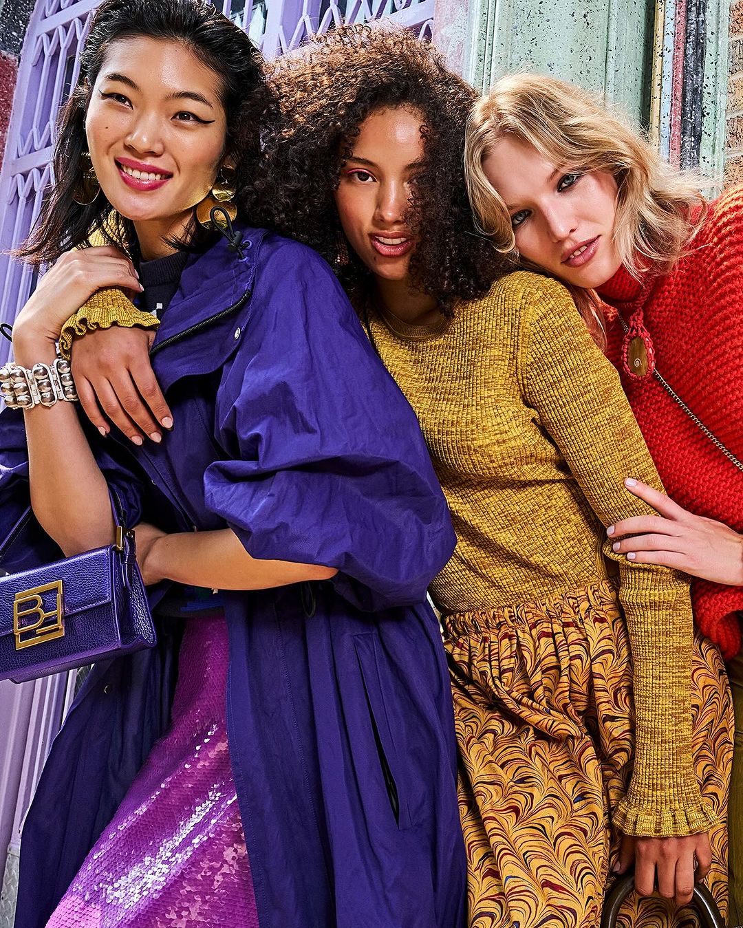 Three diverse women pose together indoors wearing fashionable clothing in red, yellow, and purple tones, with a patterned background.