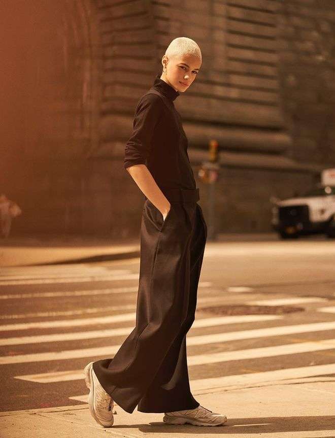 Blonde woman in black turtleneck and wide-leg trousers poses on a city crosswalk with buildings and a car in soft-focus background.