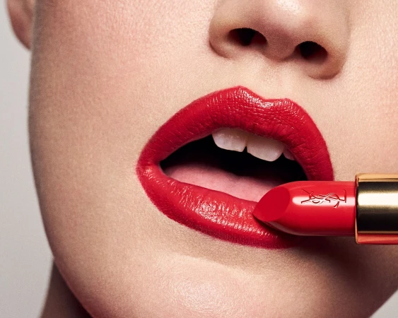 Extreme close-up of a model applying bright red lipstick with a gold tube against a plain light background