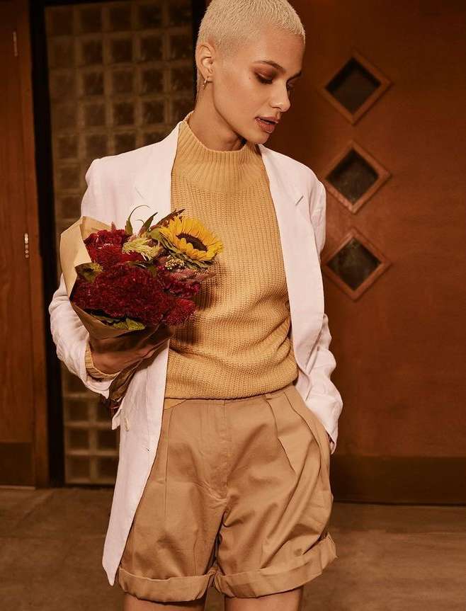Stylish woman in a white blazer, golden-brown sweater and shorts poses with flowers in an indoor setting with warm lighting.