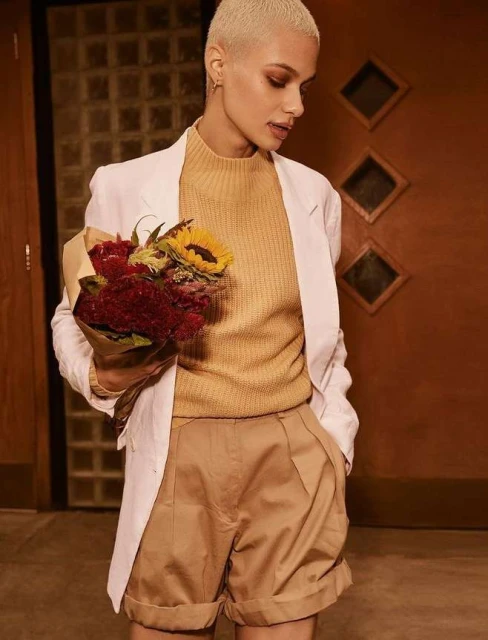 Stylish woman in a white blazer, golden-brown sweater and shorts poses with flowers in an indoor setting with warm lighting.
