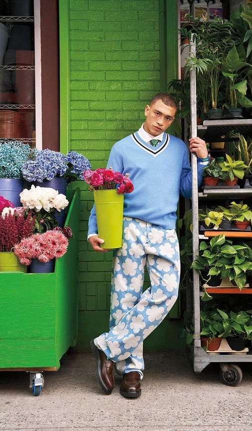 Stylish man stands by a flower shop holding a bucket of pink flowers. He is wearing patterned pants, a blue sweater, and brown dress shoes.