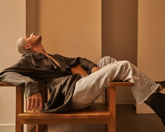 Model with short platinum blonde hair reclining in a wooden chair wearing a dark trench coat and light wash jeans