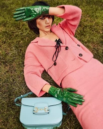 Model wearing a pink suit and glossy green gloves lying on grass with a light blue handbag