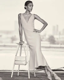 Model in a V-neck dress standing by a white stool holding a handbag, photographed by Danilo Hess.