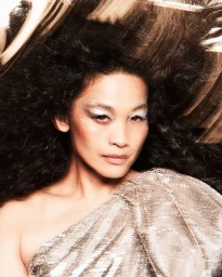 Close-up portrait of a woman with dark curly hair wearing a gold sequined top and silver glitter eye makeup by photographer Danilo Hess