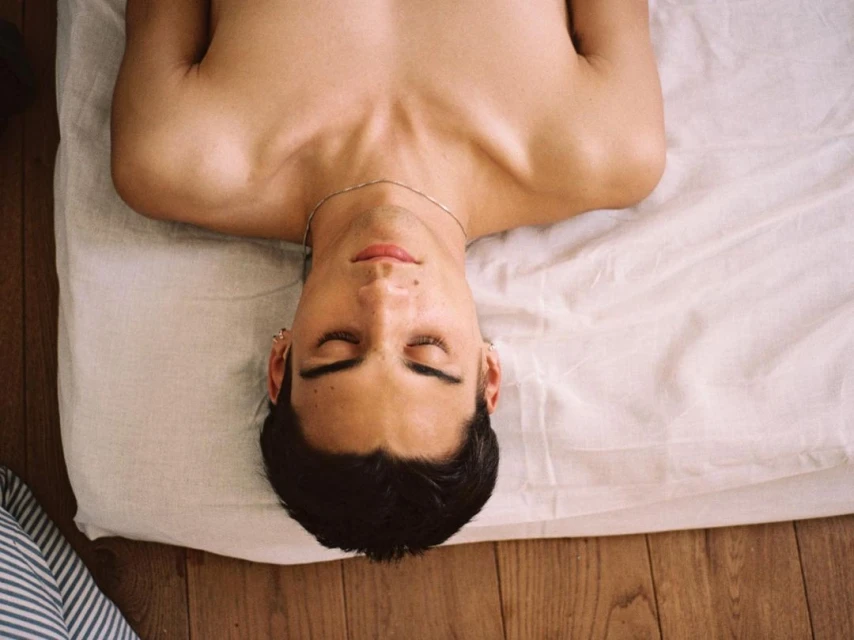 Adult male with eyes closed lays on a white sheet in a studio setting. The man is shirtless and facing the camera with a necklace.