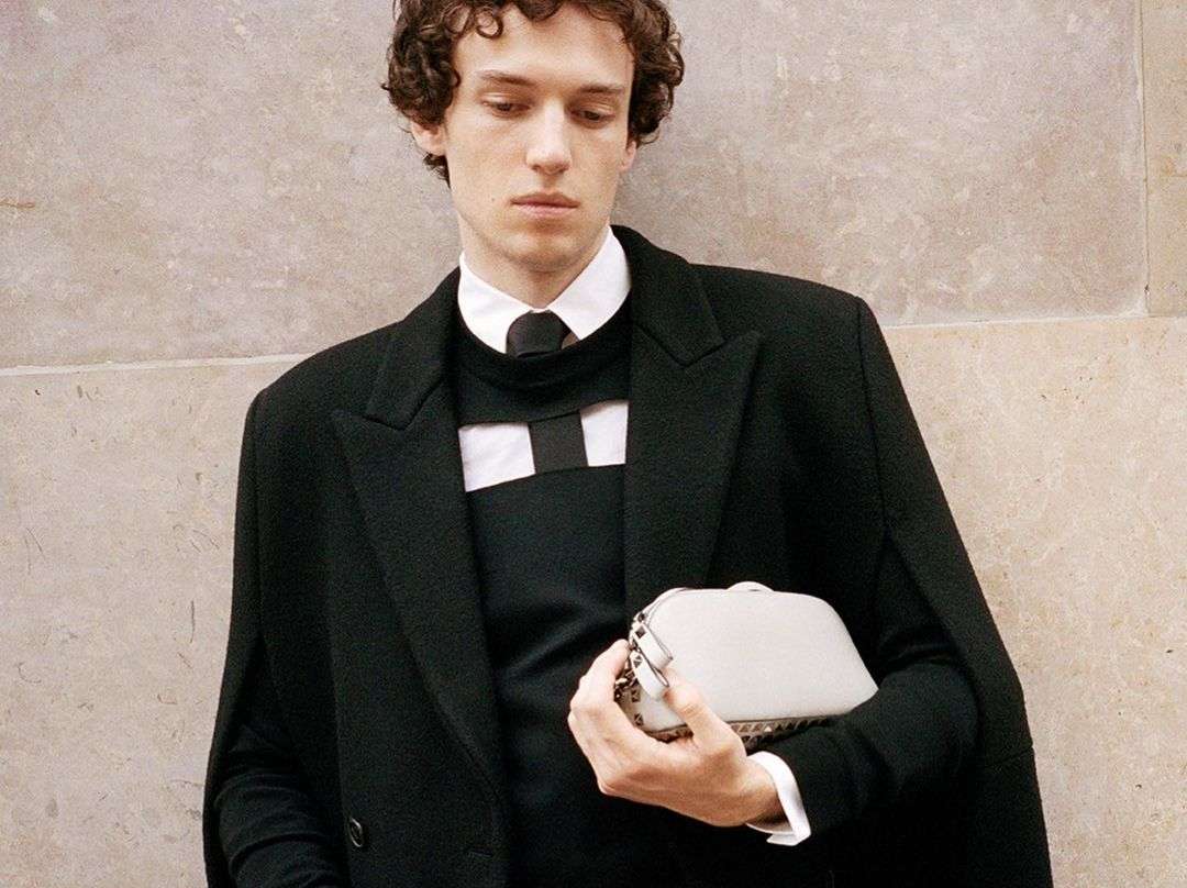 An adult male posing in a black coat and sweater with a white clutch bag in front of a light neutral textured wall indoors.