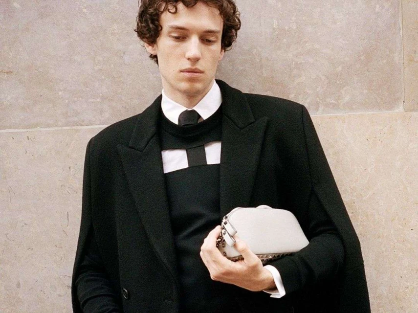 An adult male posing in a black coat and sweater with a white clutch bag in front of a light neutral textured wall indoors.