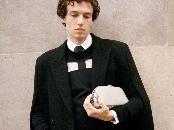 An adult male posing in a black coat and sweater with a white clutch bag in front of a light neutral textured wall indoors.