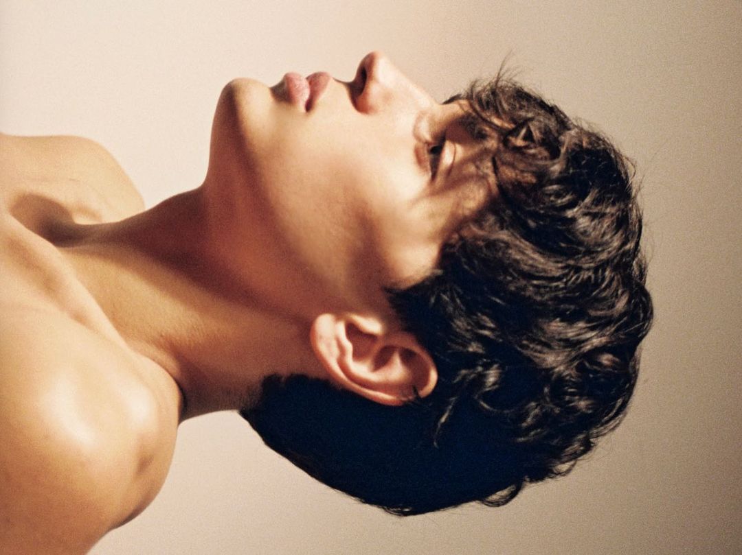 Profile portrait of a young man with dark curly hair tilted upward against a light neutral background