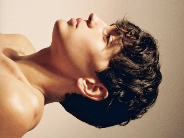 Profile portrait of a young man with dark curly hair tilted upward against a light neutral background
