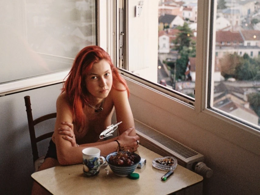 Red-haired woman sits at a table with a mug, bowl of dates, ashtray and lighter, with a view of a European town out the window.