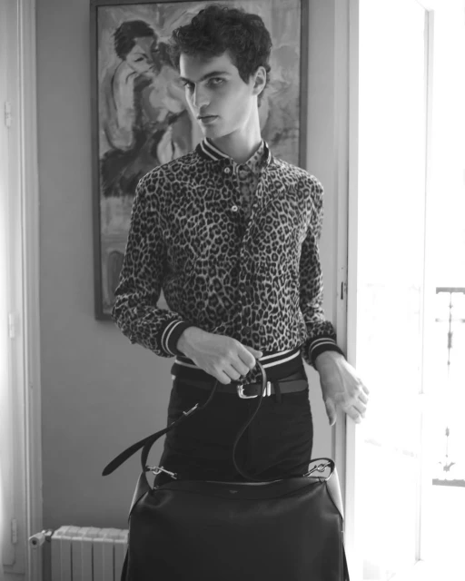 Monochrome portrait of a young adult male in patterned shirt, black pants, accessorized with a belt and black leather bag indoors.