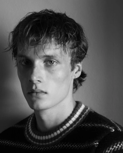Black and white portrait of a young adult male with curly hair, wearing a striped sweater, against a neutral backdrop in soft studio lighting.
