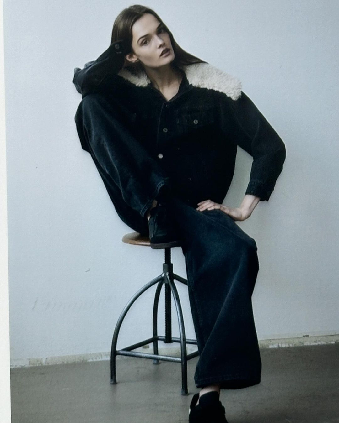 Fashion editorial shot of a female model in a dark denim jacket with fur collar and wide-leg jeans, posing on a metal stool.
