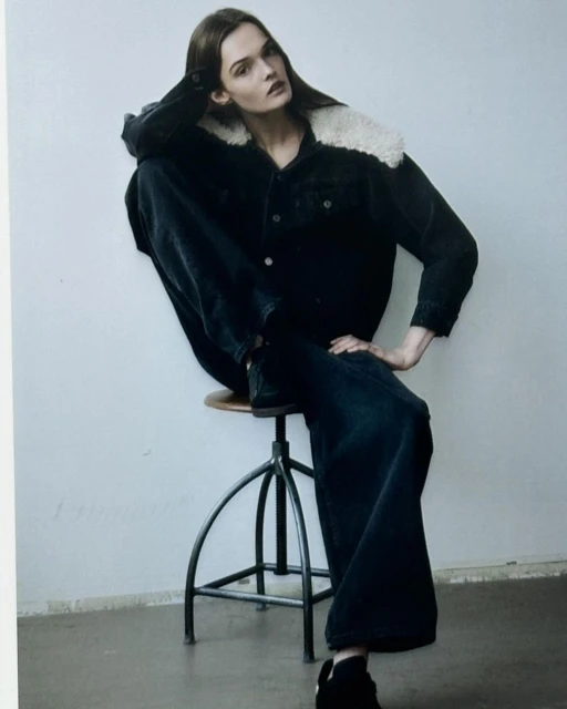 Fashion editorial shot of a female model in a dark denim jacket with fur collar and wide-leg jeans, posing on a metal stool.