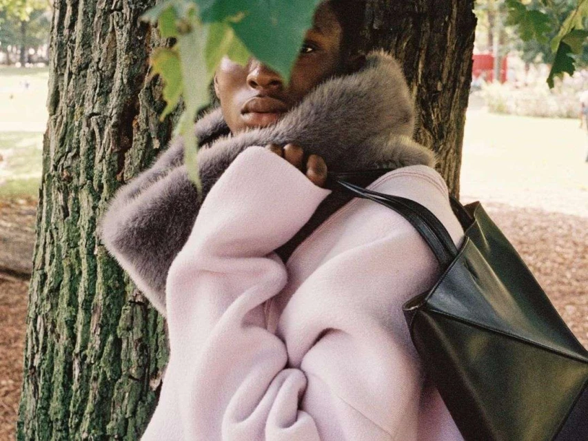 A black model posing next to a tree. The model is wearing a pink coat with a fur collar and carrying a black tote bag.