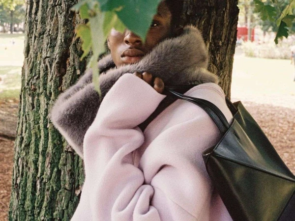 A black model posing next to a tree. The model is wearing a pink coat with a fur collar and carrying a black tote bag.