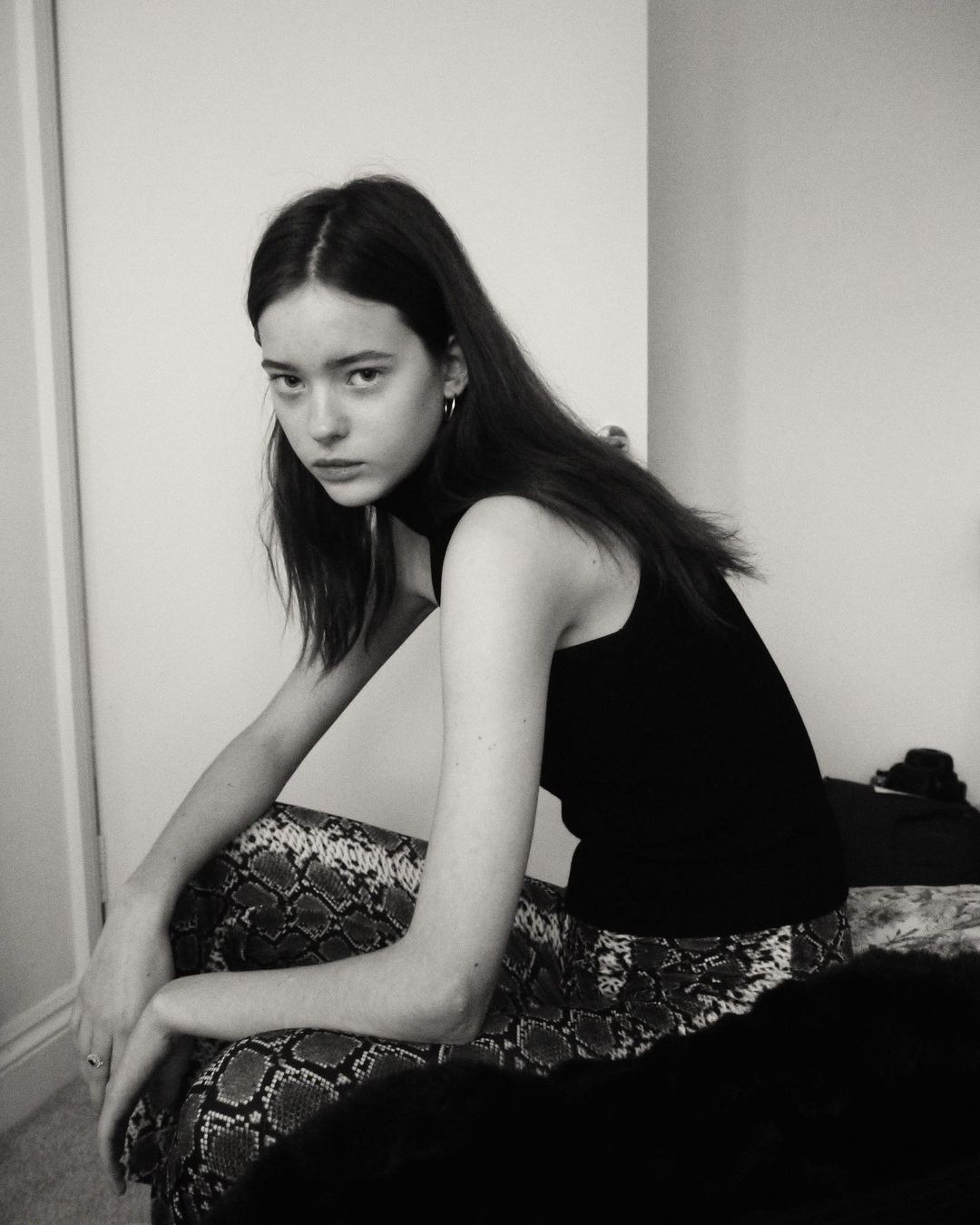 Monochrome portrait of a young woman with long brown hair, wearing a black top and patterned pants, seated indoors against a white wall.