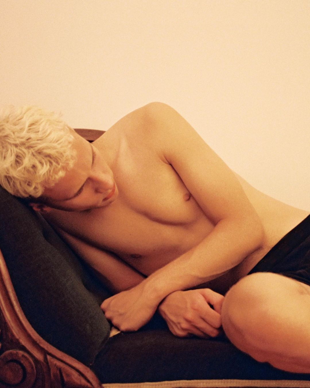 Shirtless adult male with blond hair reclines on ornate sofa in indoor setting with warm lighting, showcasing casual pose and relaxed mood.