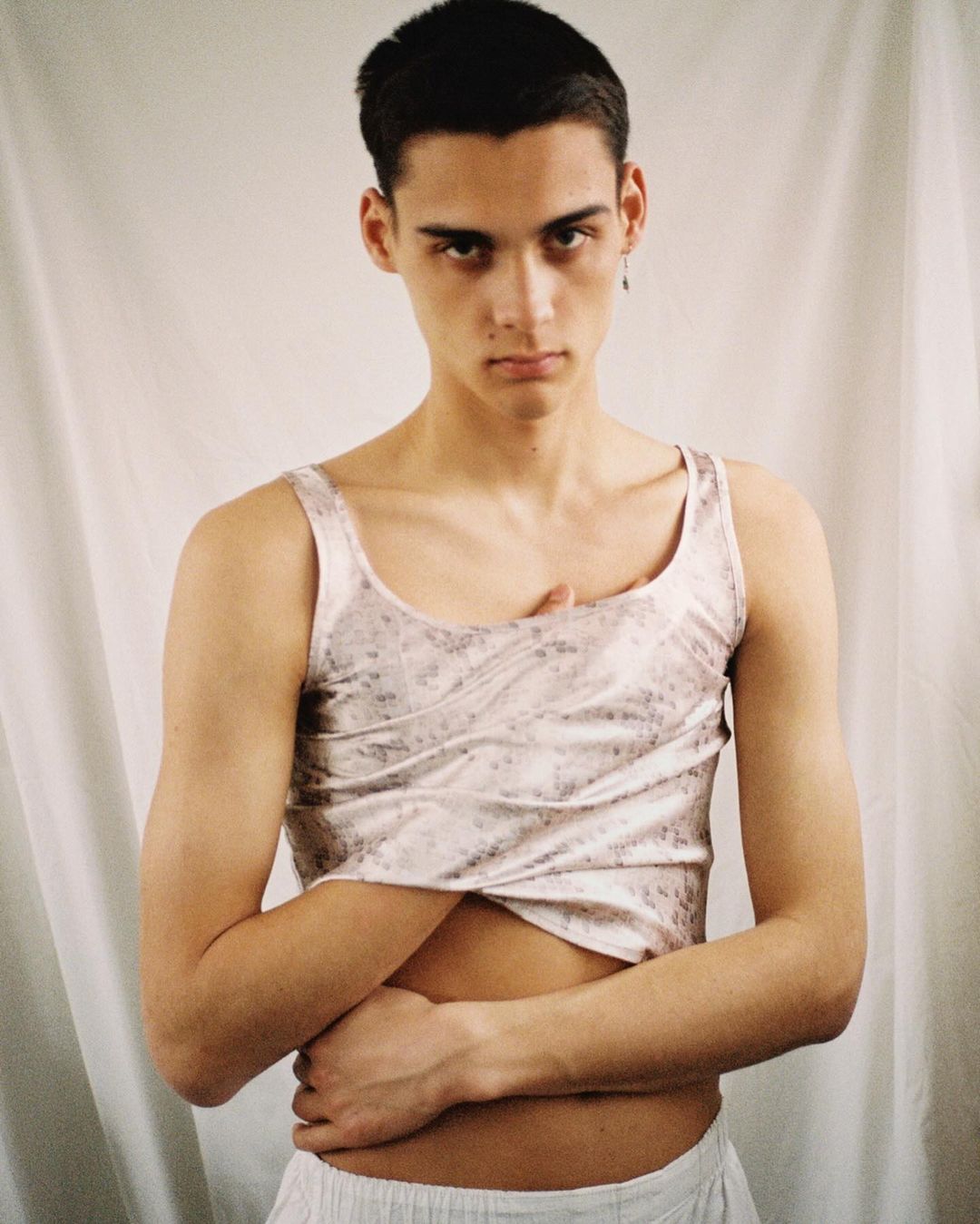 Portrait of a young adult man in a pale patterned tank top, posing indoors against a neutral backdrop with soft lighting.
