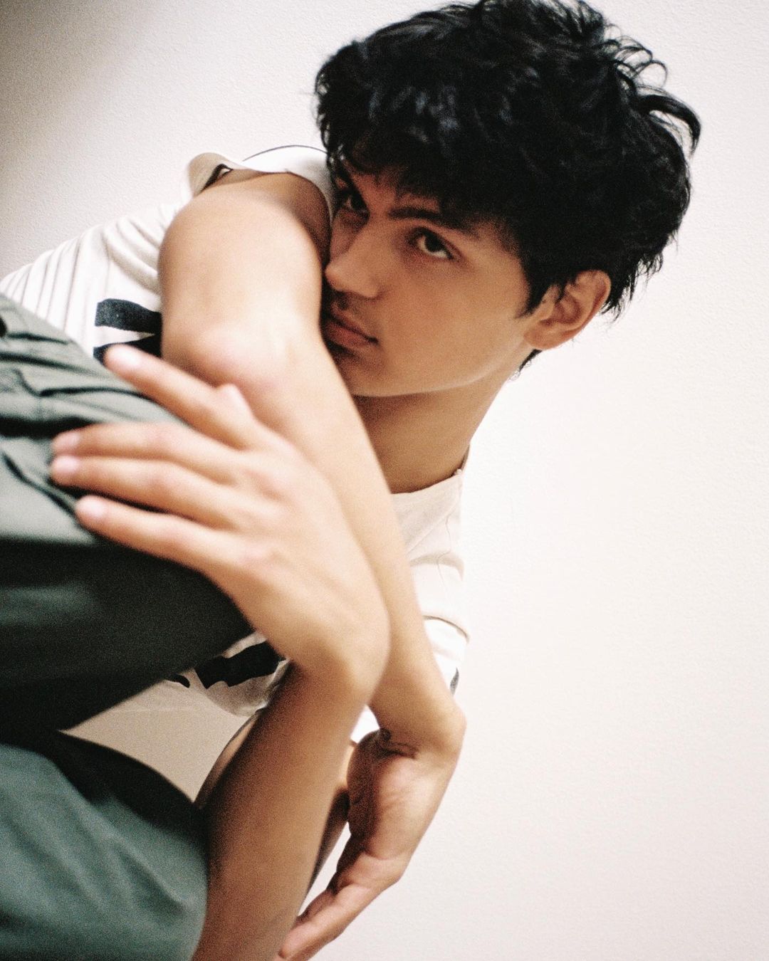 Young man posing indoors, wearing a t-shirt and green pants. Portrait-style photo, close-up, angled view, and soft, diffused lighting.