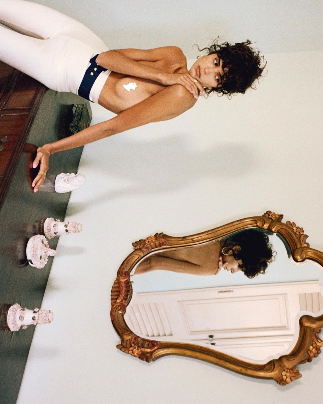 Woman poses in white pants and cream on chest near dresser and mirror indoors, with curly dark hair and jewelry on a white background.