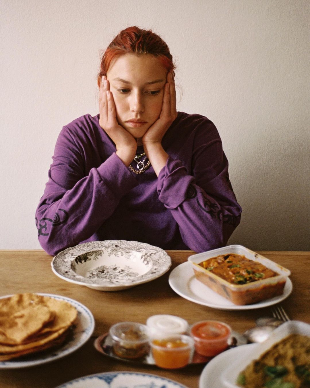 Young woman with reddish hair rests her chin on her hands at a wooden table with Indian food and various dishes in an indoor setting.