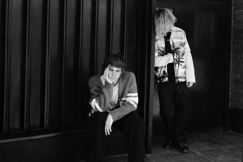 Two young men pose in front of an urban door. One is sitting with a pensive expression while the other stands next to the door.