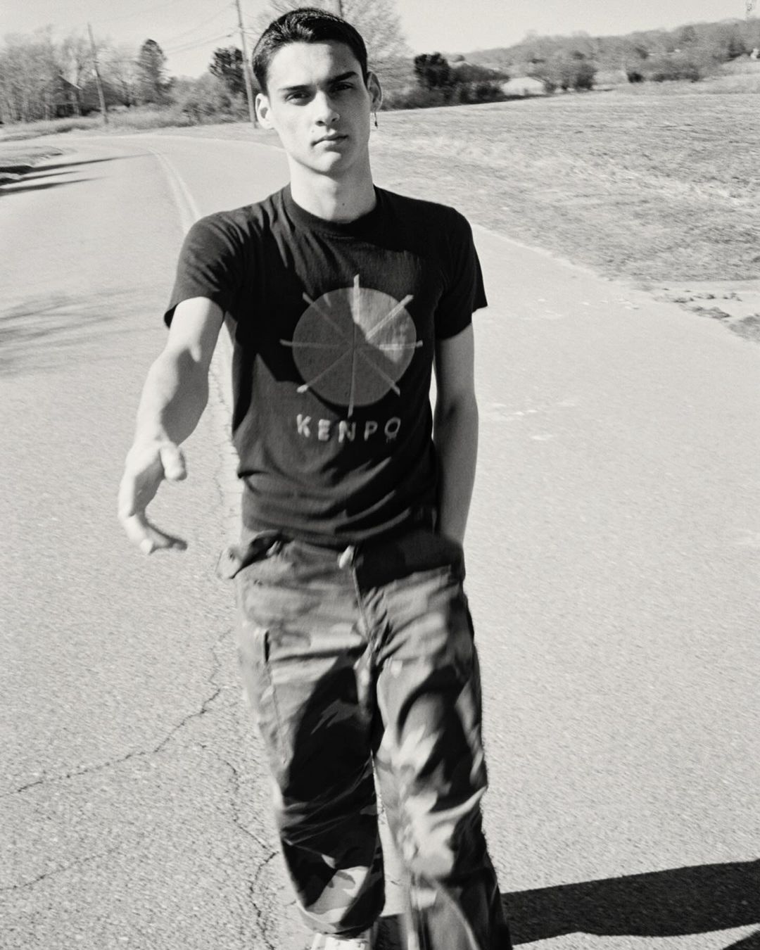 Black and white portrait of a young adult male wearing a graphic tee and camouflage pants walking outdoors on a paved road on a sunny day.